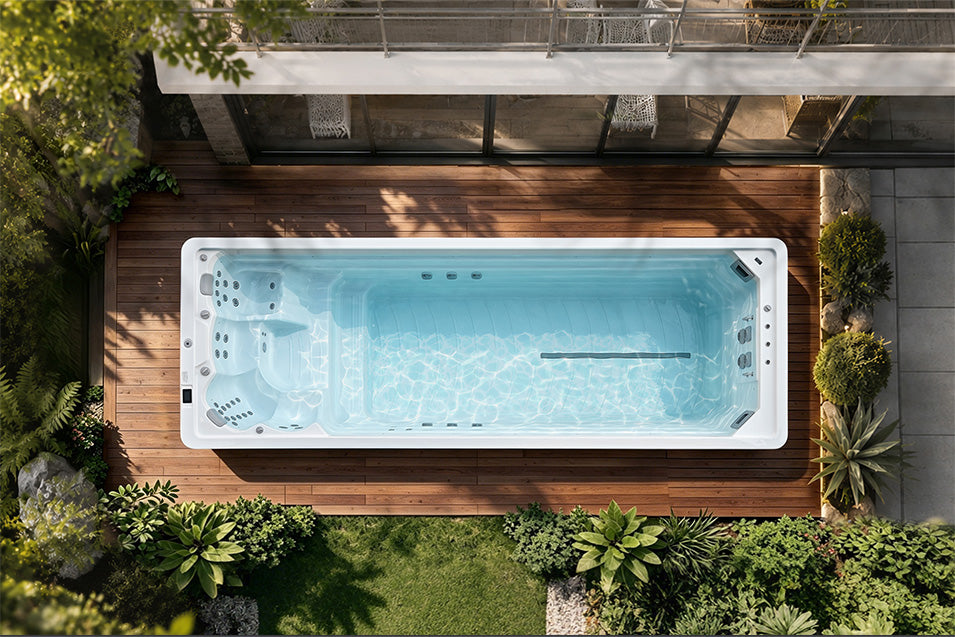 A top-down view of a rectangular outdoor swim spa with light blue water, situated on a brown wooden deck surrounded by green plants and a modern house.