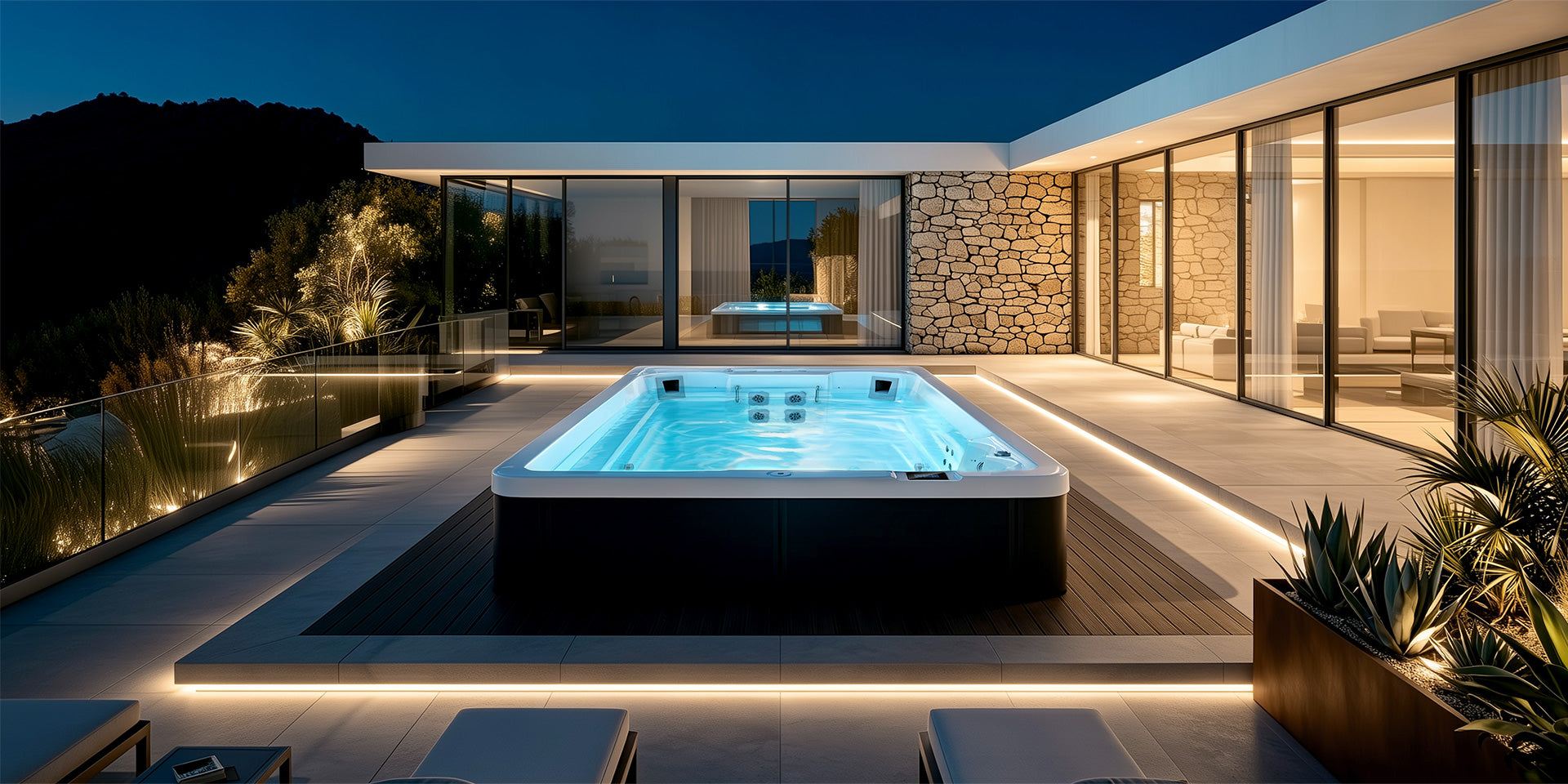 A modern outdoor swim spa with glowing blue water sits on a dark deck beside a contemporary house at night.